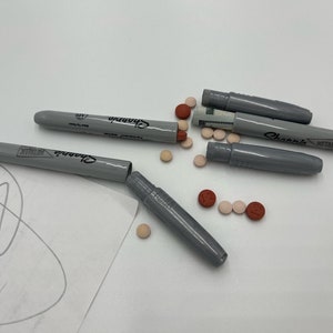 Op de afbeelding: Vier grijze Sharpie-permanent markers met de dopjes eraf, waardoor een voorraad kleine rode en roze pillen zichtbaar wordt. De markers liggen op een wit oppervlak.
