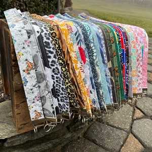 Könnte beinhalten: Eine Sammlung bunter Stoffschlüsselanhänger mit verschiedenen Mustern, darunter Tierdrucke, Blumenmuster und geometrische Formen. Die Schlüsselanhänger sind in einer Reihe angeordnet, wobei die Enden der Stoffstreifen nach unten hängen. Jeder Schlüsselanhänger hat einen silbernen Metallring am Ende.