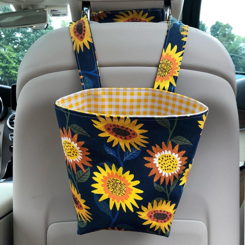 May include: A car seat organizer with a navy blue exterior featuring a sunflower pattern. The interior lining is yellow and white gingham. The organizer hangs from the headrest with matching sunflower straps.