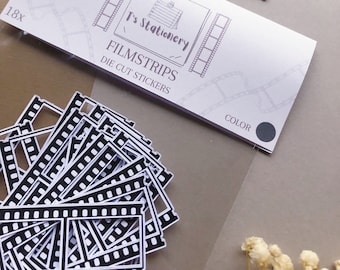 Może przedstawiać: Opakowanie 18 czarno-białych naklejek wyciętych w kształcie taśmy filmowej. Naklejki znajdują się w przezroczystym plastikowym opakowaniu z białą etykietą, na której widnieje napis "T's Stationery" i "Filmstrips Die Cut Stickers".