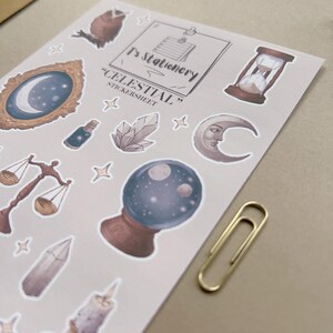 Puede incluir: Una hoja de pegatinas de tem&aacute;tica celestial con un b&uacute;ho, una luna, un reloj de arena, una balanza, un cristal y una bola de cristal. La hoja tiene el texto "T's Stationery" y "Celestial Sticker Sheet". Un clip dorado est&aacute; a la derecha.