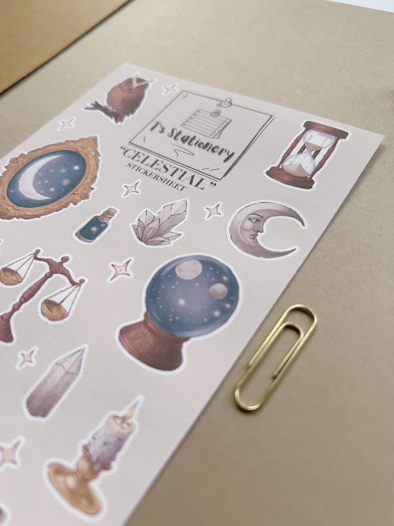 Puede incluir: Una hoja de pegatinas de tem&aacute;tica celestial con un b&uacute;ho, una luna, un reloj de arena, una balanza, un cristal y una bola de cristal. La hoja tiene el texto "T's Stationery" y "Celestial Sticker Sheet". Un clip dorado est&aacute; a la derecha.
