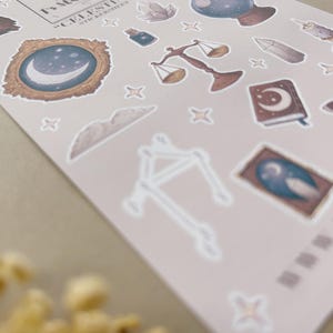 Puede incluir: Una hoja de pegatinas con dise&ntilde;os celestiales, que incluyen lunas, estrellas, cristales y balanzas. Las pegatinas tienen un estilo de acuarela con acentos dorados y azules. La hoja tambi&eacute;n incluye el texto "T's Stationery" y "Celestial Sticker Sheet".