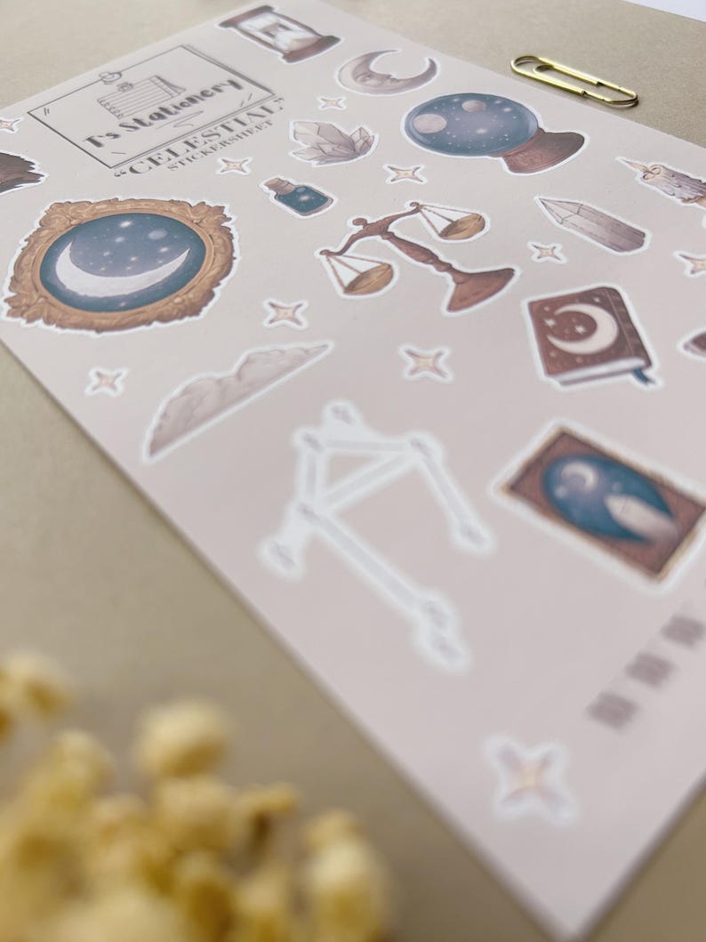 Puede incluir: Una hoja de pegatinas con dise&ntilde;os celestiales, que incluyen lunas, estrellas, cristales y balanzas. Las pegatinas tienen un estilo de acuarela con acentos dorados y azules. La hoja tambi&eacute;n incluye el texto "T's Stationery" y "Celestial Sticker Sheet".