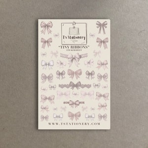 Peut inclure: Une planche d'autocollants avec des illustrations de nœuds dans des tons de rose et de blanc. Le texte "T's Stationery" et "Tiny Ribbons" sont imprimés en haut. L'adresse du site Web www.tstationery.com est imprimée en bas.