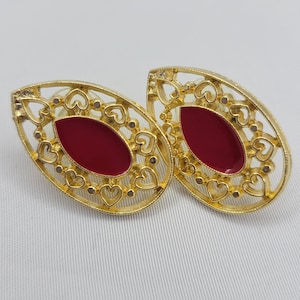 May include: A pair of gold earrings with a red oval-shaped stone set in a filigree design. The earrings are shaped like a teardrop and have a delicate, intricate pattern.