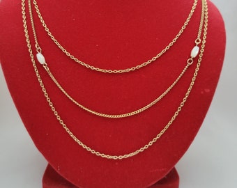 Puede incluir: Un collar dorado de varias capas con tres hilos de diferentes estilos de cadena. Cada hilo está adornado con pequeñas cuentas blancas de forma ovalada. El collar se exhibe sobre un busto de terciopelo rojo.