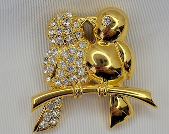 May include: A gold-toned brooch featuring two birds perched on a branch. One bird is encrusted with clear crystals, while the other is smooth. The birds are facing each other, with their beaks touching. The brooch is a decorative accessory.