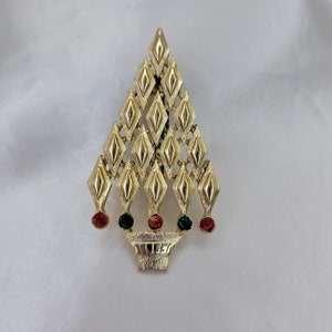May include: Gold-tone Christmas tree brooch with a basket base. The tree is made of diamond-shaped segments and adorned with red and green rhinestones. A festive accessory.