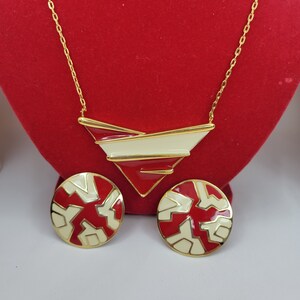 Puede incluir: Un conjunto de collar y pendientes a juego en tono dorado. El collar presenta un colgante geom&eacute;trico con detalles en rojo, crema y dorado. Los pendientes son redondos con un dise&ntilde;o similar. Las joyas se exhiben sobre un fondo de terciopelo rojo.