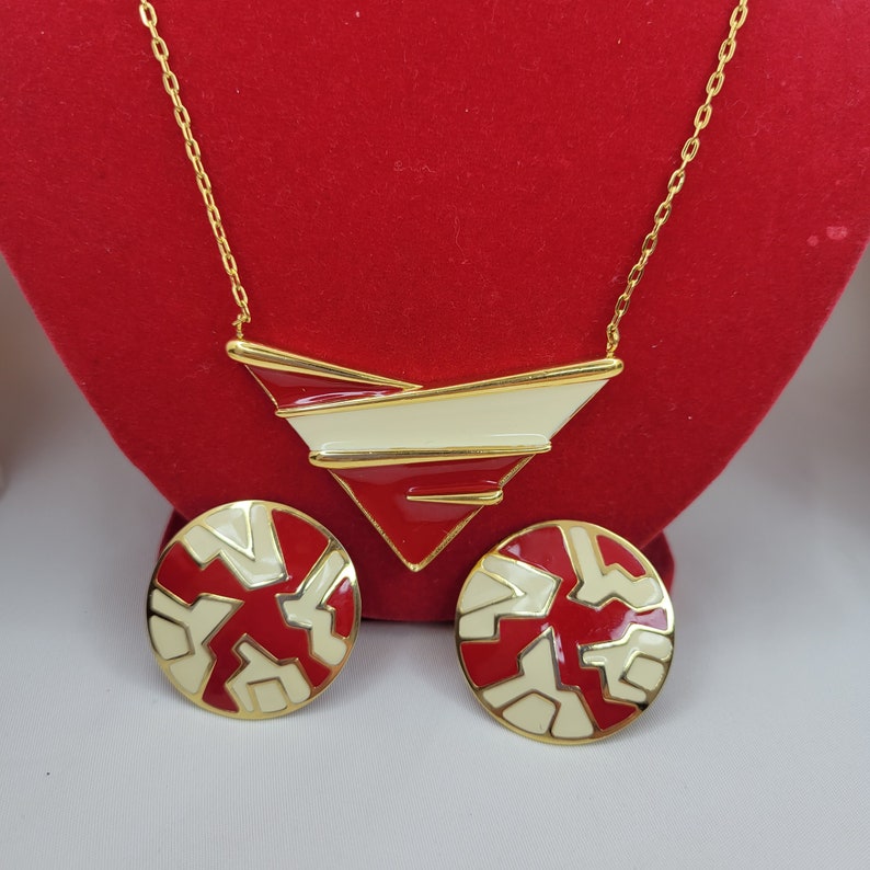 Puede incluir: Un conjunto de collar y pendientes a juego en tono dorado. El collar presenta un colgante geom&eacute;trico con detalles en rojo, crema y dorado. Los pendientes son redondos con un dise&ntilde;o similar. Las joyas se exhiben sobre un fondo de terciopelo rojo.