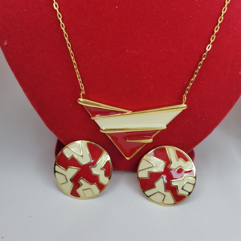 Puede incluir: Un conjunto de collar y pendientes dorados. El collar presenta un colgante geom&eacute;trico con detalles en rojo, blanco y dorado. Los pendientes son redondos con un dise&ntilde;o geom&eacute;trico similar en rojo y blanco. Las joyas se exhiben sobre una superficie roja.