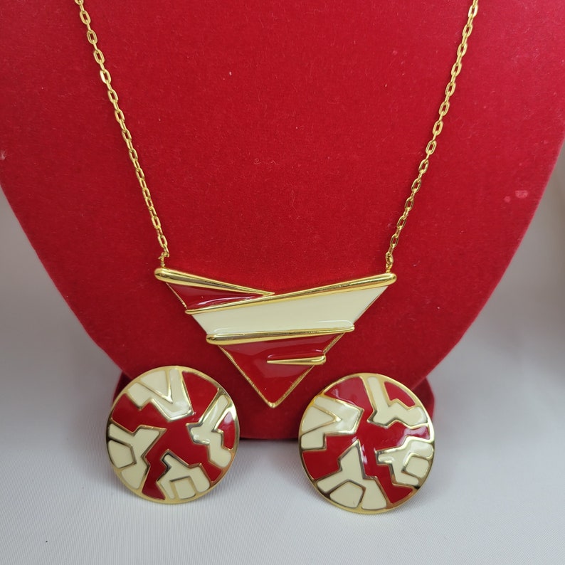 Puede incluir: Un conjunto de collar y pendientes a juego en tono dorado. El collar presenta un colgante geom&eacute;trico con detalles en rojo, crema y dorado. Los pendientes son redondos con un dise&ntilde;o similar. Las joyas se exhiben sobre una superficie roja.