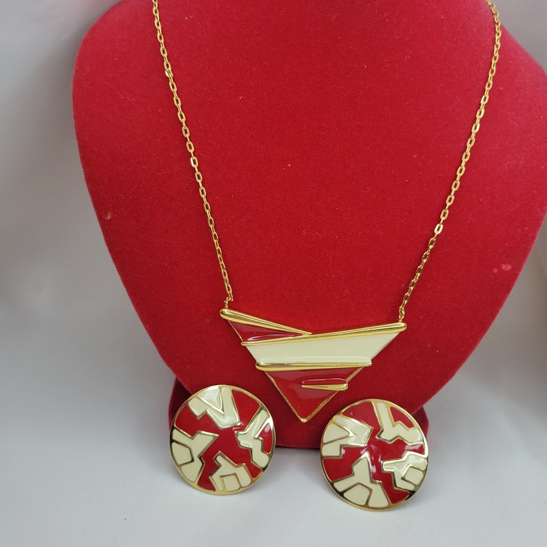 Puede incluir: Un conjunto de collar y pendientes a juego en tono dorado. El collar presenta un colgante geom&eacute;trico con esmalte rojo y crema. Los pendientes son redondos con un dise&ntilde;o similar en rojo y crema. Las joyas se exhiben sobre un fondo de terciopelo rojo.