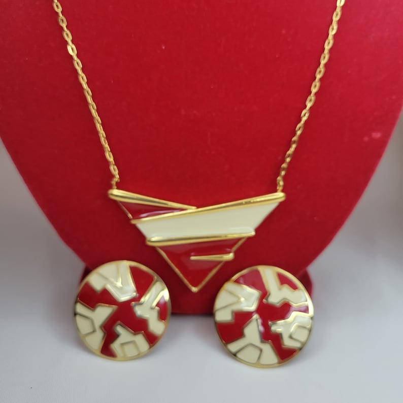 Puede incluir: Un conjunto de collar y pendientes a tono dorado. El collar presenta un colgante geom&eacute;trico con detalles en rojo, blanco y dorado. Los pendientes son redondos con un dise&ntilde;o geom&eacute;trico similar en rojo y blanco. Las joyas se exhiben sobre una superficie roja.