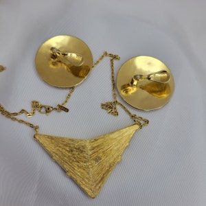 Puede incluir: Un conjunto de joyas vintage en tono dorado con un collar y pendientes de clip. El collar tiene un colgante triangular texturizado con una cadena. Los pendientes son redondos con cierres de clip. El conjunto se muestra sobre una superficie blanca.