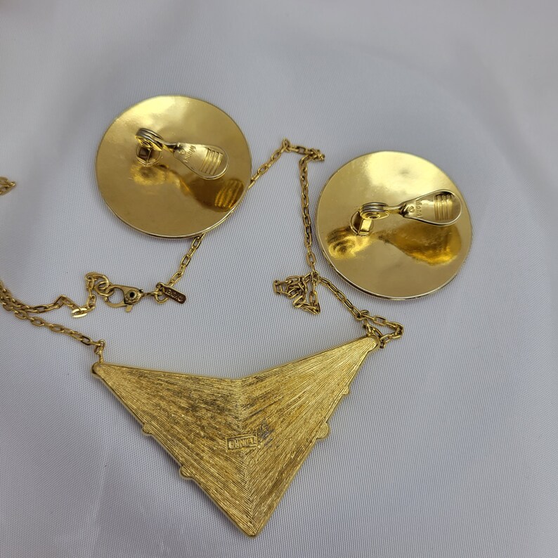 Puede incluir: Un conjunto de joyas vintage en tono dorado con un collar y pendientes de clip. El collar tiene un colgante triangular texturizado con una cadena. Los pendientes son redondos con cierres de clip. El conjunto se muestra sobre una superficie blanca.