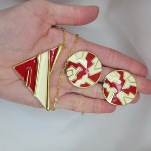 Puede incluir: Un conjunto de joyas vintage con un collar y pendientes a juego. El collar tiene un colgante geom&eacute;trico con esmalte rojo y crema, y los pendientes son redondos con un dise&ntilde;o similar. Todas las piezas tienen detalles met&aacute;licos dorados.