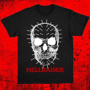 May include: Black t-shirt featuring a detailed, grayscale skull design with pins and the word "HELLRAISER" in red. The skull has large eye sockets and a detailed jawline. The shirt is a classic crew neck.