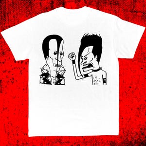 May include: White t-shirt featuring a black and white graphic of two cartoon characters. One character has a black hairstyle and is clenching a fist. The other character has a black hairstyle and is wearing a black choker.