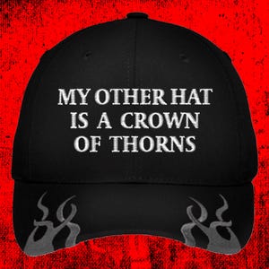May include: Black baseball cap with the text "MY OTHER HAT IS A CROWN OF THORNS" in white. The cap has a curved brim and a decorative design on the sides. The background is red.