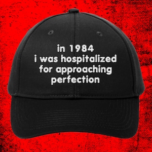 May include: Black baseball cap with white embroidered text: "in 1984 i was hospitalized for approaching perfection." The cap has a curved brim and a button on top. The hat is a classic, casual accessory.