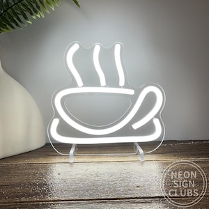 May include: A white neon coffee cup sign with steam rising from it. The sign is on a clear stand, and the text "NEON SIGN CLUBS" is visible. The sign is placed on a wooden surface.