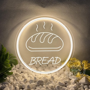 May include: White neon sign with a bread illustration and the word "BREAD" in a circle. The sign is on a bed of white rocks.