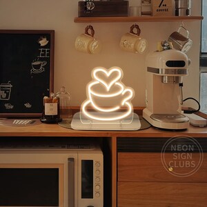 May include: A neon sign depicting a coffee cup with a heart design, emitting a warm white glow. The sign is placed on a white base. The background includes a microwave, coffee machine, and wooden shelving with mugs.