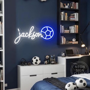 May include: White neon sign with the name "Jackson" and a blue soccer ball. The sign is hanging on a dark blue wall in a bedroom with a white dresser, a bed with blue bedding, and a desk with a laptop.