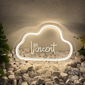 May include: White neon sign in the shape of a cloud with the name "Vincent" written in cursive inside. The sign is sitting on a bed of white rocks.