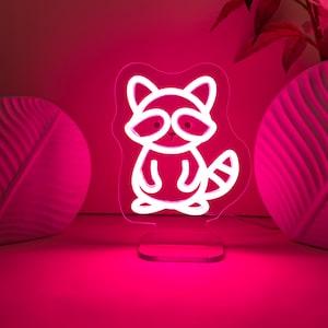 May include: A neon light in the shape of a raccoon, with a white outline and a pink glow. The raccoon has a striped tail and is standing on a clear base. The background is a solid pink color, with decorative vases and flowers.