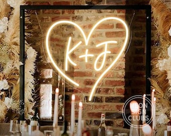 Op de afbeelding: Een neonbord in de vorm van een hart met de initialen "K + J". Het bord is omgeven door een zwart frame en geplaatst tegen een bakstenen muur. De scène omvat kaarsen, bloemen en een tafelsetting, wat duidt op een bruiloft of evenement.