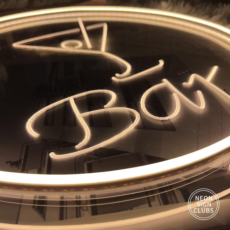 May include: A neon sign with the word "Bar" in a stylized font, with a martini glass icon above. The sign is illuminated with a warm, white light against a dark background. The sign is circular and has a white border.