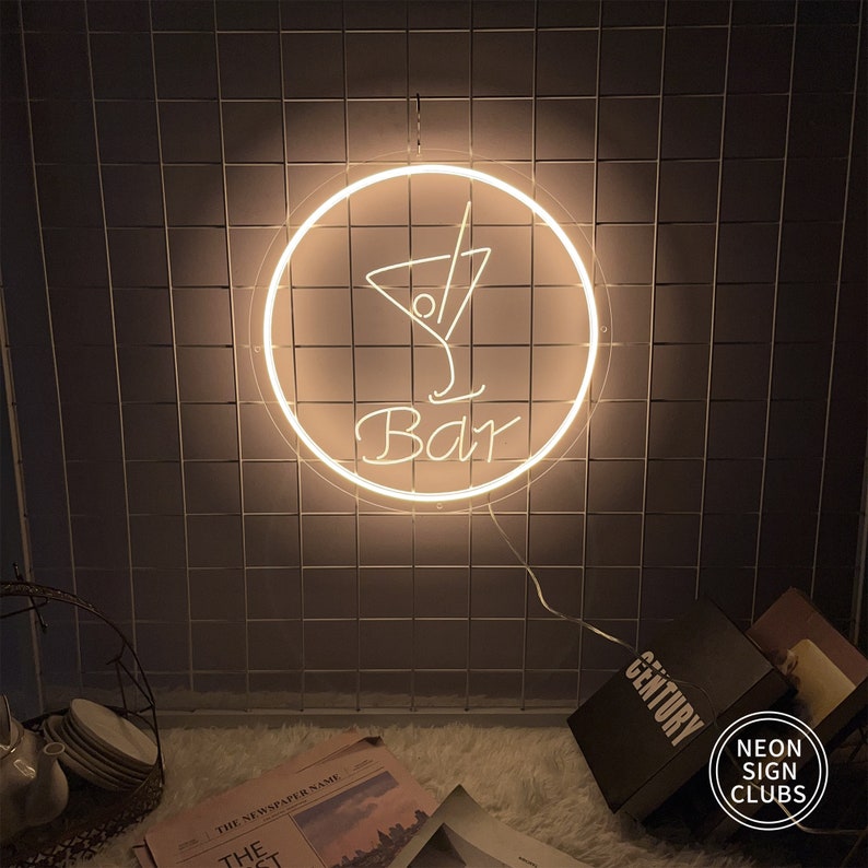 May include: A glowing neon sign with a cocktail glass design inside a circle, with the word "Bar" underneath. The sign is lit with a warm, inviting light and is mounted on a dark, tiled wall. The sign is from Neon Sign Clubs.