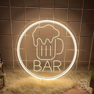 May include: A round, illuminated neon sign with a white outline. The sign features a beer mug graphic above the word "BAR" in a bold, sans-serif font. The sign is designed for a bar or pub setting.