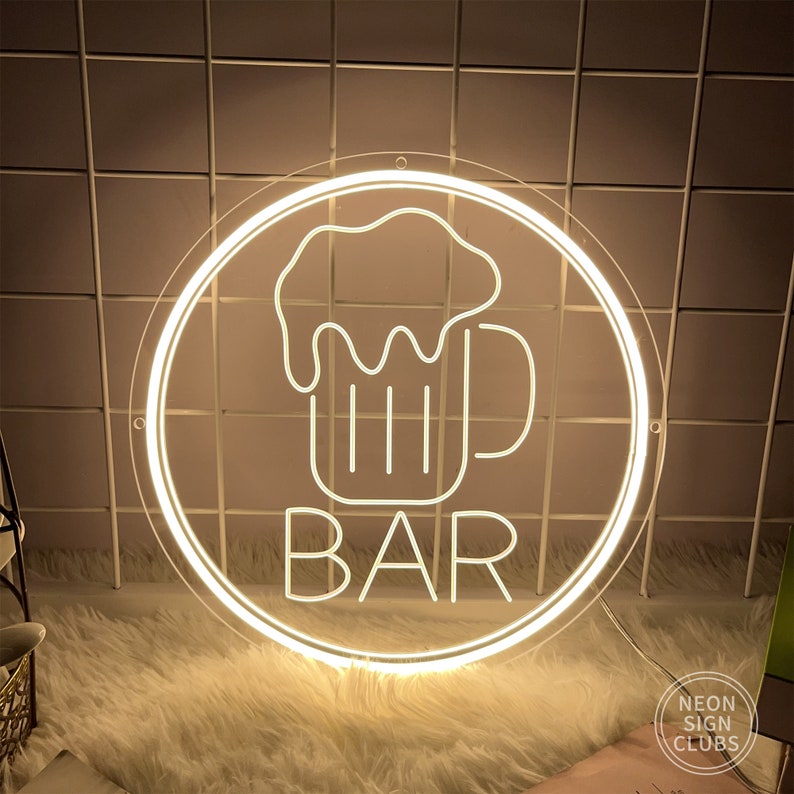 May include: A round, illuminated neon sign with a white outline. The sign features a beer mug graphic above the word "BAR" in a bold, sans-serif font. The sign is designed for a bar or pub setting.