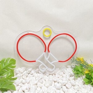 May include: A neon sign in the shape of two red ping pong paddles and a yellow ball. The paddles are connected by white lines. The sign is mounted on a clear acrylic base. The background is white with white stones and greenery.