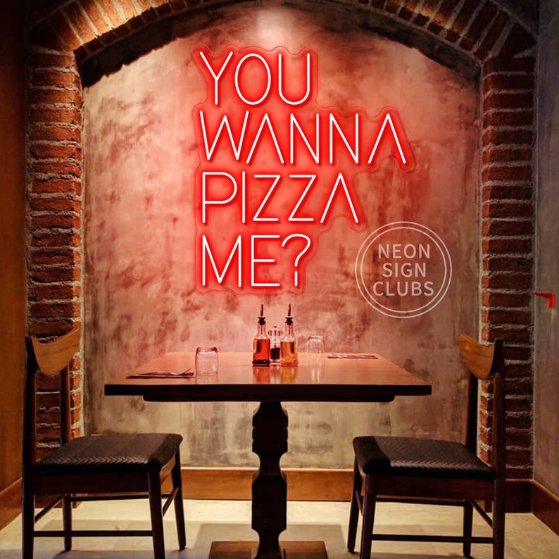 May include: A bright red neon sign mounted on a textured wall displaying the text YOU WANNA PIZZA ME? in a stylized font. The sign is positioned above a wooden table with two chairs, featuring a circular logo with the text NEON SIGN CLUBS.