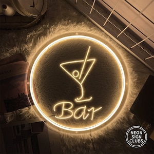 May include: A round neon sign with a cocktail glass design and the word "Bar" in a cursive font. The sign is illuminated with a warm, yellow glow and is surrounded by a circular frame. The sign is on a fluffy, white surface.