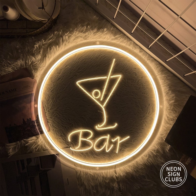 May include: A round neon sign with a cocktail glass design and the word "Bar" in a cursive font. The sign is illuminated with a warm, yellow glow and is surrounded by a circular frame. The sign is on a fluffy, white surface.