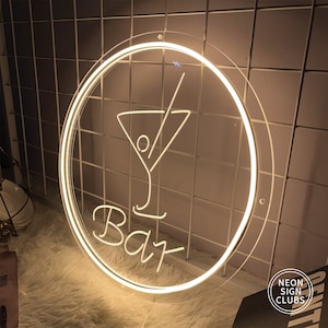 May include: A round, illuminated neon sign with a cocktail glass design and the word "Bar" in script. The sign has a clear border and is lit with a warm white light. The sign is mounted on a white grid.