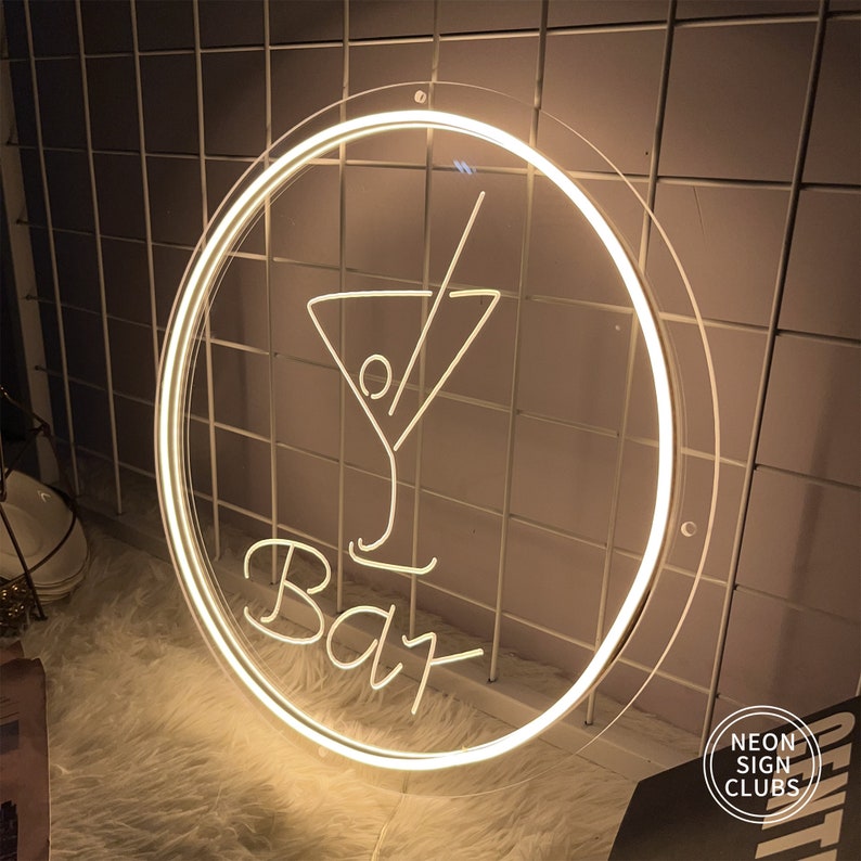 May include: A round, illuminated neon sign with a cocktail glass design and the word "Bar" in script. The sign has a clear border and is lit with a warm white light. The sign is mounted on a white grid.