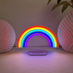 Puede incluir: Una lámpara de neón con forma de arcoíris en colores vibrantes: rojo, naranja, amarillo, verde, azul y morado. La luz se apoya sobre una base rectangular transparente. En el fondo hay jarrones decorativos y vegetación.