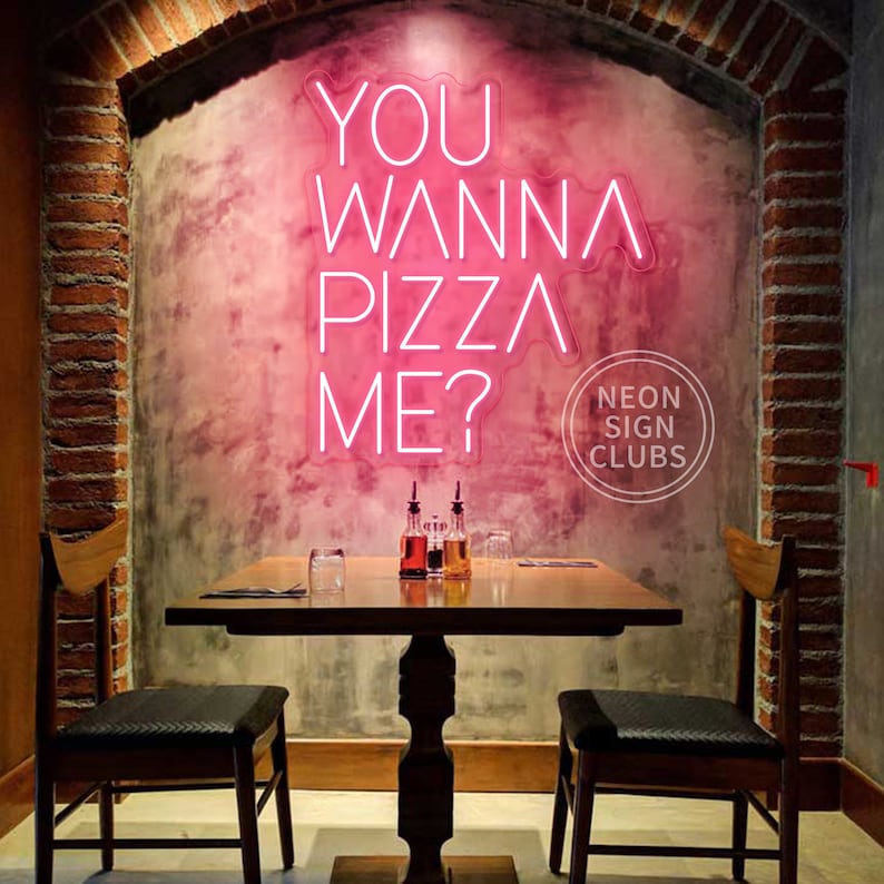 May include: A bright pink neon sign mounted on a textured wall displaying the text YOU WANNA PIZZA ME? in a modern sans-serif font. The sign is positioned above a wooden dining table with two chairs, featuring a circular logo in the corner.