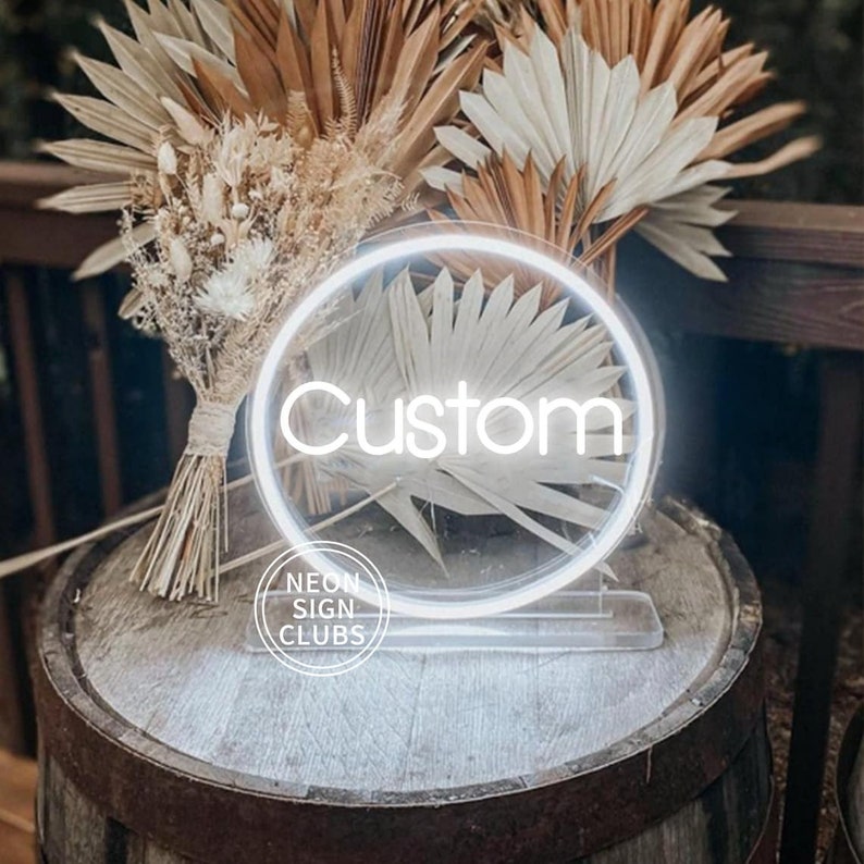 May include: A white neon sign with the word "Custom" inside a circular frame. The sign is on a clear stand, placed on a wooden barrel. Dried flowers and palm leaves are arranged in the background. The sign has the text "NEON SIGN CLUBS" at the bottom.