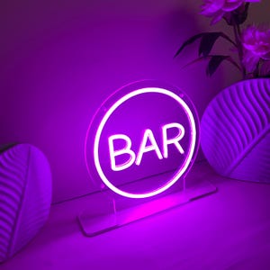 May include: A neon sign in a circular shape with the word "BAR" in white letters. The sign is lit with a bright purple light and is mounted on a clear acrylic base. The background includes a white vase with purple flowers.