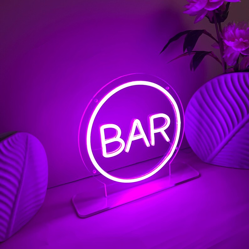 May include: A neon sign in a circular shape with the word "BAR" in white letters. The sign is lit with a bright purple light and is mounted on a clear acrylic base. The background includes a white vase with purple flowers.