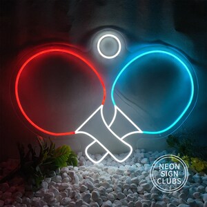 May include: Neon sign featuring two ping pong paddles, one red and one blue, with a white ball above. The sign is set against a dark background and the text "NEON SIGN CLUBS" is visible.