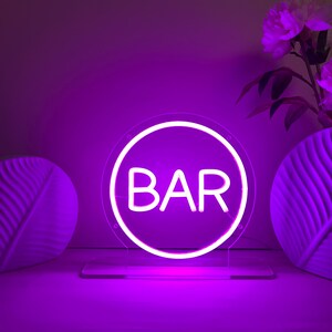 May include: A neon sign with the word "BAR" in white letters inside a white circular frame. The sign is illuminated with a bright purple light and sits on a clear stand. Decorative vases and flowers are in the background.
